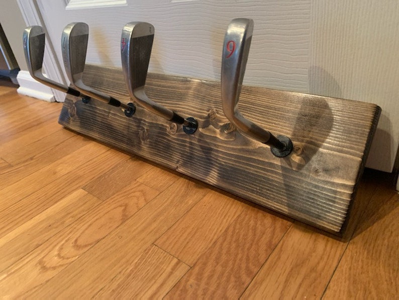 May include: A wooden wall-mounted coat rack with five golf club hooks. The rack is stained dark brown and has a rustic look.