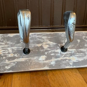 May include: A rustic wooden rack with four golf club heads mounted on it. The rack is painted white with a distressed finish. The rack has a small tag attached to it that says "Old Golf Club".