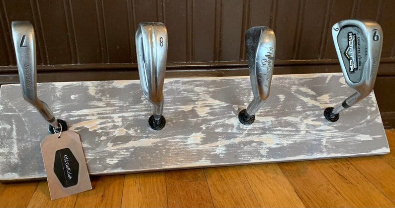 May include: A rustic wooden rack with four golf club heads mounted on it. The rack is painted white with a distressed finish. The rack has a small tag attached to it that says "Old Golf Club".