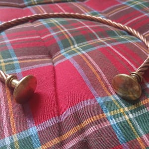 Puede incluir: Un collar torque de metal en tono cobre con una banda trenzada similar a una cuerda. Los dos extremos del collar terminan en discos planos y lisos de color latón, creando un diseño abierto que se curva en forma de semicírculo.