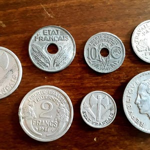 May include: A collection of vintage French coins, featuring various designs and denominations. The coins are silver-colored and display inscriptions such as "ETAT FRANCAIS" and "LIBERTE EGALITE FRATERNITE". The coins are arranged on a wooden surface.