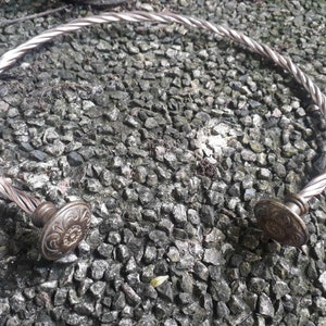 Może przedstawiać: Srebrny naszyjnik choker z drutu skręconego z dwoma ozdobnymi, okrągłymi zakończeniami. Zakończenia mają kwiatowy wzór.
