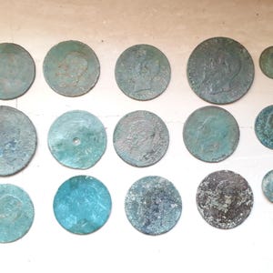 Puede incluir: Una colección de 14 monedas antiguas de cobre con varios diseños y pátina. Las monedas están dispuestas en un patrón aleatorio sobre una superficie beige claro.