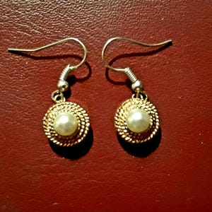 Puede incluir: Un par de pendientes dorados con una perla central y un diseño de cuerda trenzada alrededor. Tienen cierres de gancho sencillos. El fondo es de color rojo oscuro.