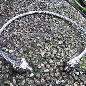 May include: Silver twisted wire bracelet with wolf head ends. The bracelet is on a gray and green background.