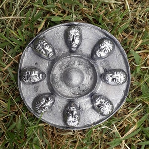 Puede incluir: Un plato circular de plata con ocho caras en relieve alrededor del borde. Las caras son todas diferentes y tienen un aspecto ligeramente desgastado.