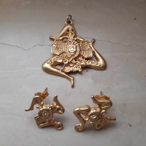 May include: A gold-tone pendant and earrings set featuring the Trinacria symbol, a three-legged figure with a head in the center. The pendant has a loop for attaching a chain, while the earrings have post backs.