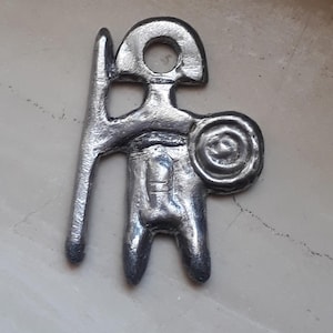 May include: A silver metal pendant depicting a stylised figure holding a spear and shield. The figure has a round head with a hole at the top for hanging.