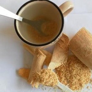 Op de afbeelding: Een witte keramische mok met een bruine rand en een houten vijzel en stamper. De mok is gevuld met een lichtbruin poeder en er zit een lepel in de mok. De vijzel en stamper zijn gevuld met een lichtbruin poeder. Er ligt een hoop lichtbruin poeder op de tafel.