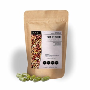 May include: A brown paper bag of "True Selenium" dietary supplement capsules with a white label and a mix of nuts and seeds. Six green capsules are scattered in front of the bag. The label includes text about supporting thyroid, antioxidant, and memory.