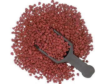 May include: A pile of small, reddish-brown annatto seeds with a dark gray scoop. The seeds are densely packed, filling the frame against a white background. The scoop is positioned in the center, partially submerged in the seeds.