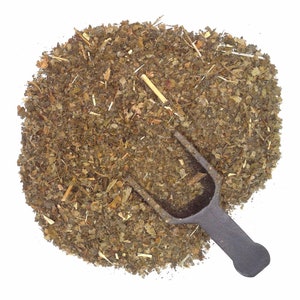 May include: A pile of dried green herbs with a small, dark gray scoop. The herbs are a mix of green and brown, with visible stems and leaf fragments. The scoop is made of metal and is positioned in the herbs.