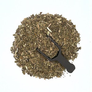 May include: A close-up of a pile of dried green herbs with a black plastic scoop. The herbs are loose and appear to be finely chopped.