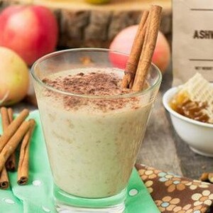 May include: A glass of creamy, light-coloured smoothie topped with cinnamon and cocoa powder. The smoothie is surrounded by apples, cinnamon sticks, and honey in a bowl. The image also includes a bag of "Ashwagandha" powder.
