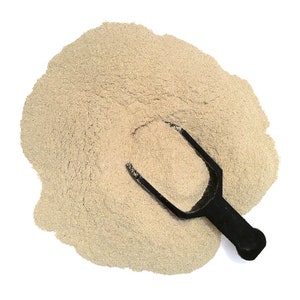 May include: A scoop of beige powder on a white background. A black plastic scoop is partially submerged in the powder.