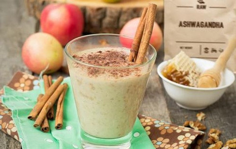 May include: A glass of creamy, light-colored smoothie topped with cinnamon and cocoa powder. The smoothie is surrounded by apples, cinnamon sticks, and honey.