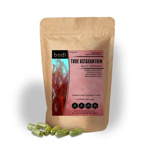May include: A brown paper bag of True Astaxanthin health supplement capsules with the brand name "bodi". The bag has a pink label with text including "Powerful Antioxidant" and "Improve Brain Health". Several green capsules are scattered in front of the bag.