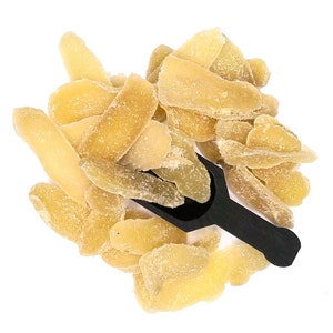 May include: A close-up of a pile of dried, candied ginger pieces. The ginger is a light yellow color and has a slightly translucent appearance. A black scoop is partially visible in the pile of ginger.