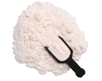 May include: A large, irregular pile of fine, white powdery substance with a textured, clumpy appearance. A small, black plastic scoop is partially submerged into the center of the mound, filled with a portion of the white powder.