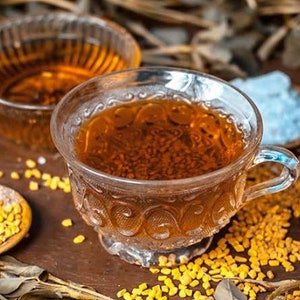 May include: A glass cup filled with a brown liquid, surrounded by yellow seeds and a small bowl of brown powder. The cup has a decorative pattern and a handle.