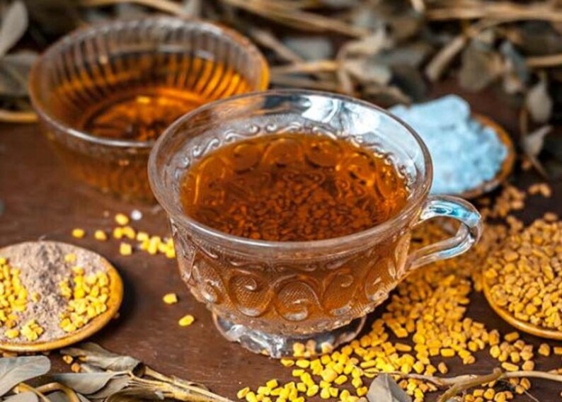 May include: A glass cup filled with a brown liquid, surrounded by yellow seeds and a small bowl of brown powder. The cup has a decorative pattern and a handle.