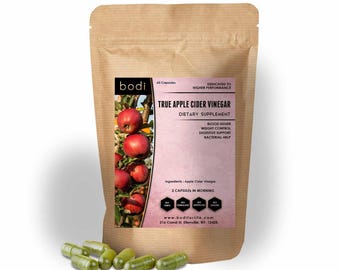 May include: A brown paper bag of "True Apple Cider Vinegar" dietary supplement capsules. The bag has a pink label with text and an image of red apples. Several green capsules are scattered in front of the bag.