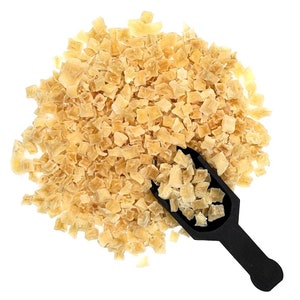 May include: A heap of diced, dried pineapple pieces, pale yellow in colour. A black scoop holds some of the pineapple. The image is taken from above against a white background.