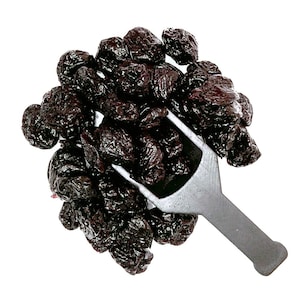 May include: A close-up of a metal scoop filled with dried prunes. The prunes are dark brown and wrinkled.