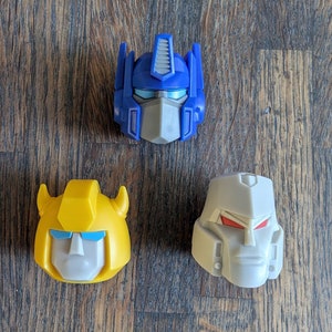 Op de afbeelding: Drie plastic speelgoed robotkoppen. De blauwe kop is Optimus Prime, de gele kop is Bumblebee en de grijze kop is Megatron.