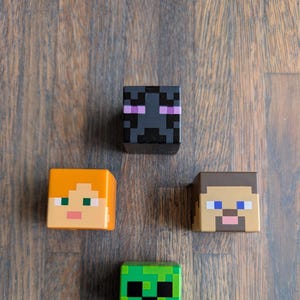 Könnte beinhalten: Vier Minecraft-Charakter-Figuren. Die Figuren sind aus Kunststoff und etwa 5 cm groß. Die Charaktere sind Steve, Alex, Creeper und Enderman. Die Figuren befinden sich auf einer Holzoberfläche.
