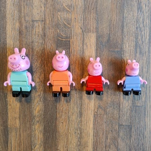Op de afbeelding: Vier roze varkensfiguurtjes met verschillende gekleurde kleding. Het varken links draagt lichtblauw, de volgende draagt oranje, de volgende draagt rood en de laatste draagt blauw.