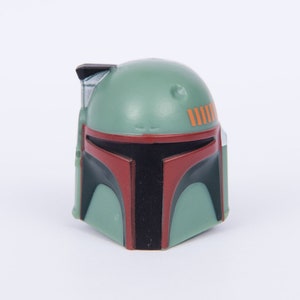Op de afbeelding: Een groen en rood plastic Boba Fett helm speelgoed. De helm heeft een zwart vizier en oranje strepen.