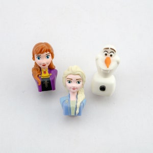 Könnte beinhalten: Drei Charakterfiguren aus dem Animationsfilm Frozen. Die Figuren umfassen Anna mit rotbraunem Haar und einem lila Kleid, Elsa mit blondem Haar und einem blauen Kleid und Olaf, den Schneemann.
