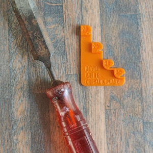 Op de afbeelding: Een beitel met een rode handgreep, een roestig metalen blad en een oranje kunststof maatlat met de nummers 20, 25, 30, 35, 40, 45, 50 en 55. De maatlat heeft de tekst "FINAL CUT" en "GLUE UP" erop gedrukt.