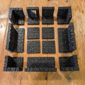 Op de afbeelding: Een verzameling zwarte, getextureerde miniatuur kerker terreinstukken. De set bevat muren, hoeken en vloertegels, allemaal ontworpen om op bakstenen te lijken. De stukken zijn gerangschikt op een houten oppervlak.