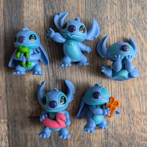 Op de afbeelding: Vijf blauwe Stitch-figuurtjes uit de Disney-film Lilo & Stitch. Elk figuurtje heeft een andere uitdrukking en houdt een ander accessoire vast. De accessoires zijn een groene kikker, een roze opblaasbare flamingo en een rood-oranje cadeaudoos.