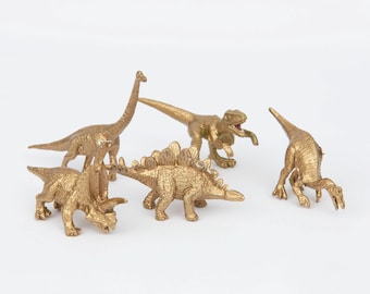 May include: Five gold-colored plastic dinosaur figurines. The dinosaurs include a stegosaurus, a brachiosaurus, a velociraptor, a triceratops, and a parasaurolophus.