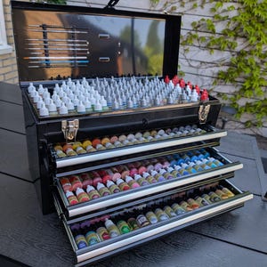 Puede incluir: Una caja de herramientas de artista de metal negro con múltiples cajones y compartimentos. La sección superior abierta contiene pinceles, mientras que los cajones están llenos de numerosas botellas de pintura pequeñas de varios colores. La caja de herramientas está sobre una superficie oscura.