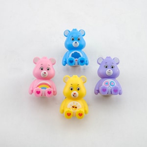 Op de afbeelding: Vier Care Bear-figuurtjes. Een blauwe Care Bear met een wolk en een ster op zijn buik, een roze Care Bear met een regenboog op zijn buik, een gele Care Bear met een zon op zijn buik en een paarse Care Bear met een hart en twee lolly's op zijn buik.
