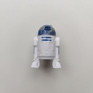 Op de afbeelding: Een witte en blauwe speelgoedrobot met een rond hoofd en een zwart oog. De robot heeft een zilveren koepel op zijn hoofd en een blauw paneel op zijn borst.