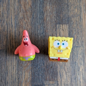 Op de afbeelding: Twee cartoonfiguren, een roze en een gele, op een houten achtergrond. Het roze personage is Patrick Star uit Spongebob Squarepants, en het gele personage is Spongebob Squarepants.