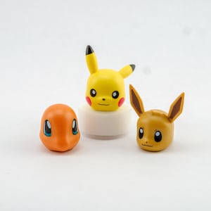Op de afbeelding: Drie kleine plastic figuurtjes van Pokémon-personages. Een gele Pikachu, een oranje Vuurvlieg en een bruine Eevee.