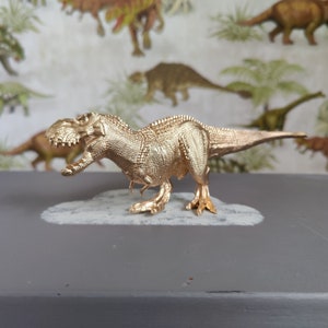 Può includere: Una statuetta di dinosauro Tyrannosaurus Rex color oro. La statuetta è in posizione eretta con la bocca aperta e la testa leggermente inclinata di lato. La statuetta è dettagliata con squame testurizzate e denti affilati.