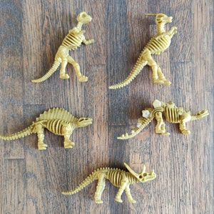 Op de afbeelding: Vijf plastic dinosaurus skeletten in verschillende poses. De skeletten zijn beige en hebben een textuur. De skeletten zijn gerangschikt op een houten oppervlak.