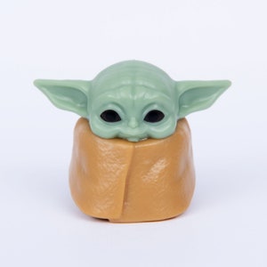 Op de afbeelding: Een klein groen en bruin beeldje van een personage uit de Star Wars-franchise, bekend als "The Child" of "Baby Yoda". Het beeldje draagt een bruine robe en heeft grote, donkere ogen.