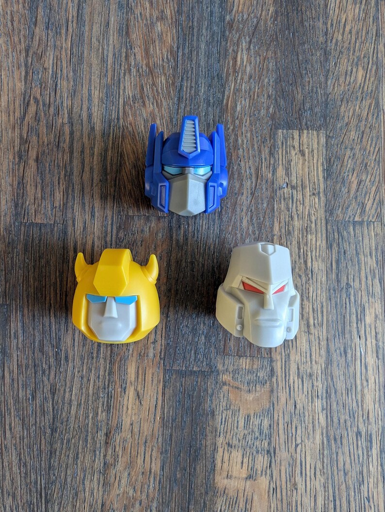 May include: Three plastic toy robot heads. The blue head is Optimus Prime, the yellow head is Bumblebee, and the grey head is Megatron.