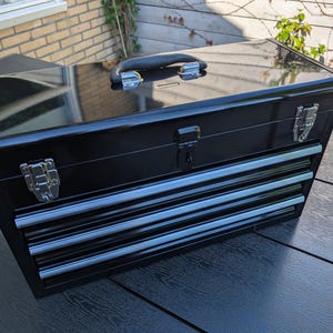 May include: A black toolbox with three drawers and a top compartment. The toolbox has a shiny finish, a black handle, and silver latches. The drawers have silver accents. The toolbox is designed for storing tools and equipment.