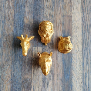 Puede incluir: Cuatro figuritas de cabeza de animal de color dorado. Un león, una jirafa, un hipopótamo y un rinoceronte.