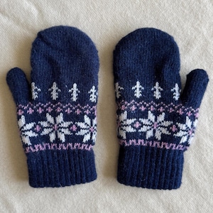 Könnte beinhalten: Ein Paar dunkelblaue Strickfäustlinge mit einem weißen und hellrosa Fair-Isle-Muster. Das Design zeigt eine Reihe weißer Schneeflockenmotive in der Mitte, die von dünnen rosa Linien und kleinen weißen Lilien-Symbolen darüber begrenzt werden.