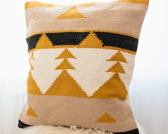 May include: A decorative pillow with a geometric design in beige, black, and mustard yellow. The pillow is rectangular and rests on a fluffy, cream-colored stool with wooden legs. The pillow's pattern includes triangles and horizontal stripes.